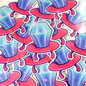 May include: A pile of colorful stickers featuring a cartoon ring with a large, iridescent blue gem. The ring is set on a pink base with a white outline. The stickers are perfect for adding a touch of whimsy to any project.