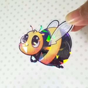 Può includere: Un adesivo olografico di un'ape di cartone animato con occhi grandi e un corpo a strisce gialle e nere. L'ape ha ali iridescenti.