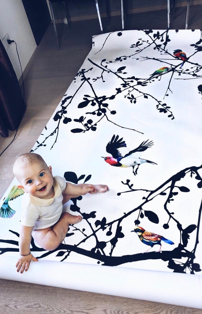 May include: A baby sits on a white and black wallpaper roll with a black silhouette of branches and colorful birds. The wallpaper has a repeating pattern of branches and birds.