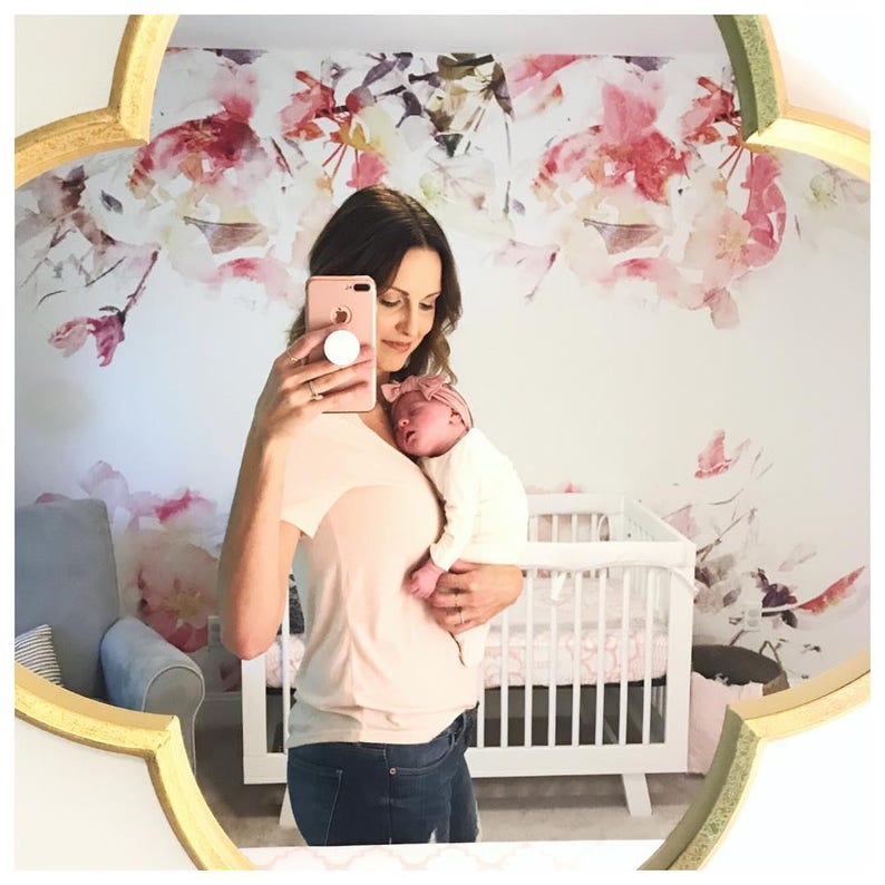 May include: A woman in a pink shirt and blue jeans holds a baby in her arms. The baby is wearing a white onesie and a pink headband. The woman is taking a selfie with a pink phone. The background is a white wall with a floral wallpaper.