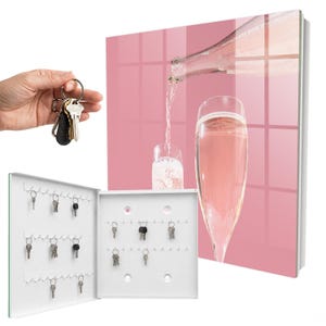 May include: A white metal key cabinet featuring a pink glass front panel with a printed image of a bottle pouring sparkling pink liquid into a glass. The interior includes multiple rows of hooks for organising keys, with several keys hanging.