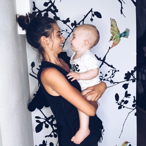 May include: A woman in a black tank top and denim shorts holds a baby in her arms. The baby is wearing a white shirt with the word "Love" on it. The background is a white wall with black silhouettes of branches and birds.