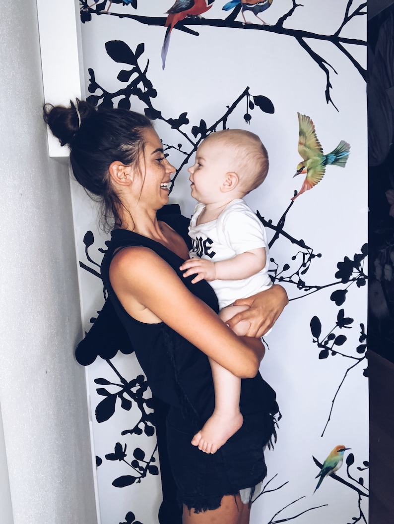 May include: A woman in a black tank top and denim shorts holds a baby in her arms. The baby is wearing a white shirt with the word "Love" on it. The background is a white wall with black silhouettes of branches and birds.
