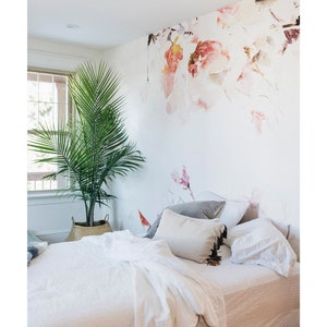 May include: A white bed with a white duvet and pillows, a grey pillow, and a brown and white pillow with tassels. The bed is in a bedroom with a white wall and a floral wallpaper. A green plant is in a wicker basket on the floor.
