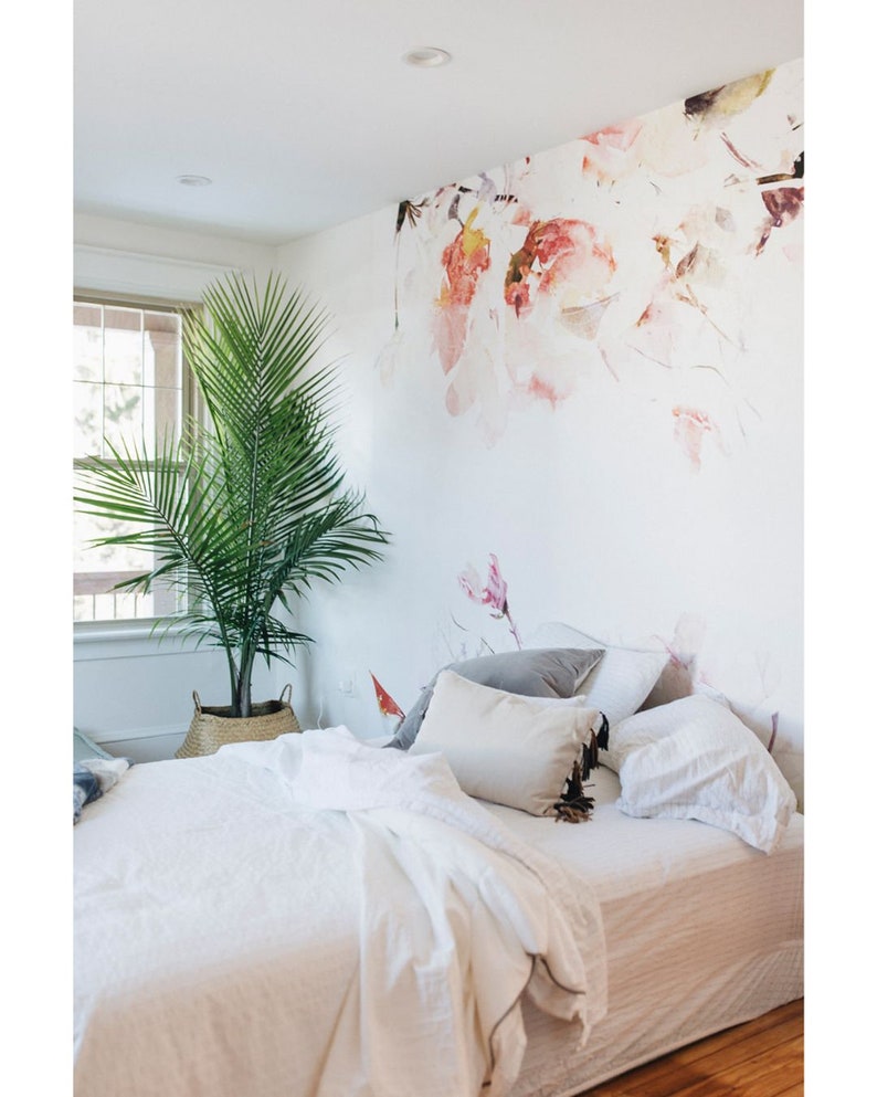 May include: A white bed with a white duvet and pillows, a grey pillow, and a brown and white pillow with tassels. The bed is in a bedroom with a white wall and a floral wallpaper. A green plant is in a wicker basket on the floor.