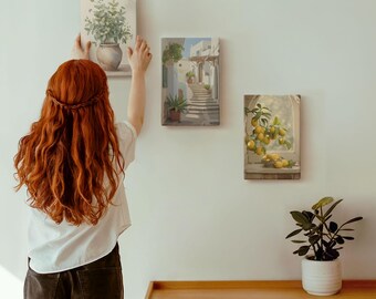 May include: A person hanging three canvas art prints on a white wall. The prints depict a vase of greenery, a street scene with stairs, and lemons on a branch. A wooden cabinet and a potted plant are in the foreground.