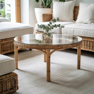 May include: A circular coffee table featuring a clear glass top and a natural rattan base. The table has four legs and a woven design. A small glass vase with green leaves sits on the table. The table is part of a living room set, likely in a conservatory.
