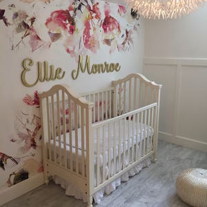 May include: A white crib with a white ruffled skirt and a white floral wallpaper with the name "Ellie Monroe" in gold letters.