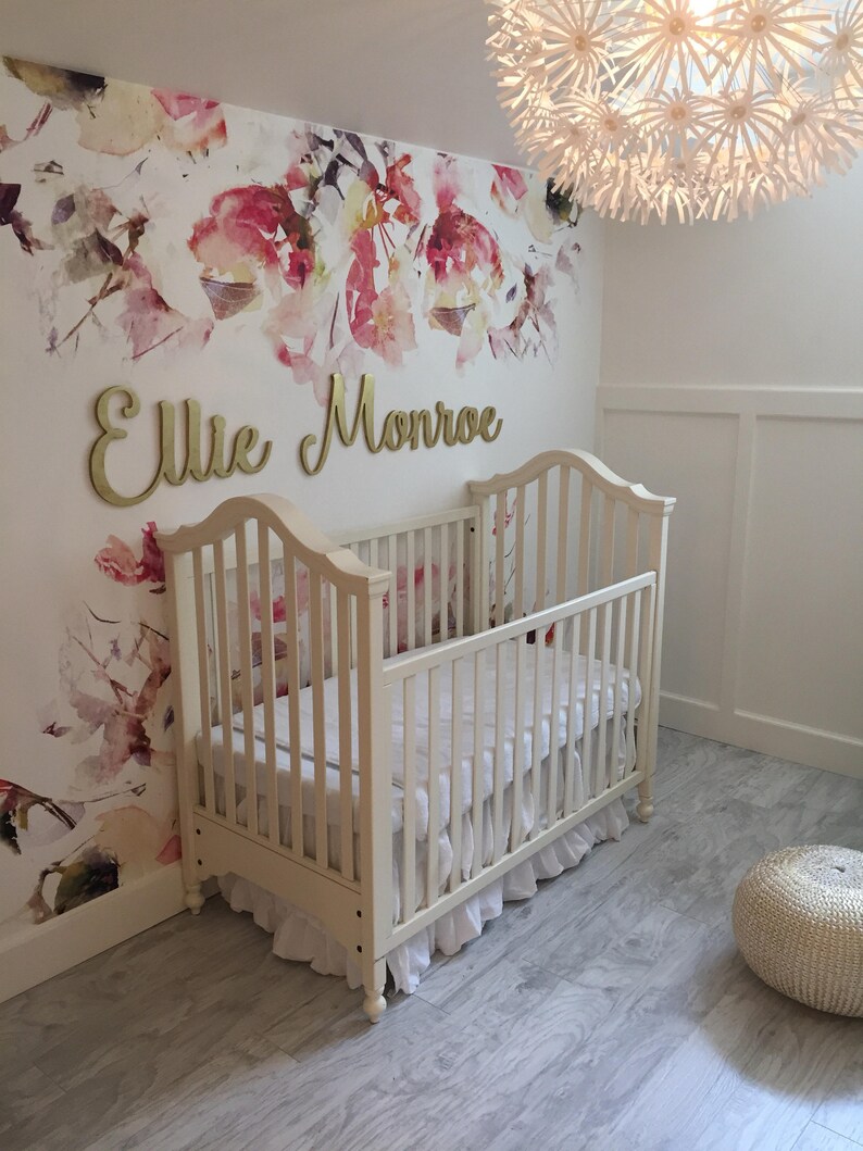 May include: A white crib with a white ruffled skirt and a white floral wallpaper with the name "Ellie Monroe" in gold letters.