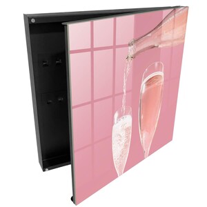 Op de afbeelding: Een zwarte sleutelkast voor aan de muur met een scharnierende deur voorzien van een roze grafische print van een fles die mousserende wijn in twee hoge glazen schenkt. De binnenkant is zwart en bevat vier kleine metalen haken voor sleutels.