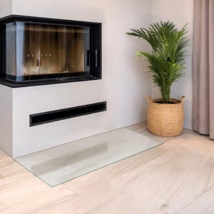 Könnte beinhalten: Ein moderner Eckkamin mit schwarzem Rahmen und Glasfront sowie einem rechteckigen schwarzen Lüftungsschlitz darunter. Eine grüne Palme steht in einem geflochtenen beigen Korb neben dem Kamin auf einem hellen Holzboden.