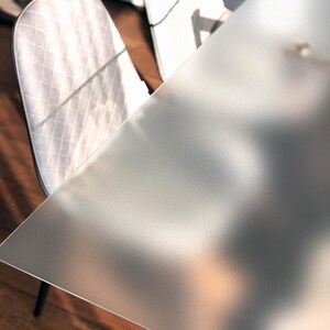 Puede incluir: Una mesa de comedor moderna con una superficie de vidrio esmerilado. Se ve parte de una silla blanca acolchada. El suelo de madera añade calidez a la escena. La superficie de la mesa refleja la luz, creando un brillo suave y difuso. La mesa mide unos 120 cm de largo.