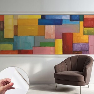 Op de afbeelding: Abstracte kunstprint met een geometrisch ontwerp van kleurrijke rechthoekige vormen in geel, blauw, rood en roze. Het kunstwerk is ingelijst en wordt tentoongesteld boven een bruine fauteuil met metalen poten.