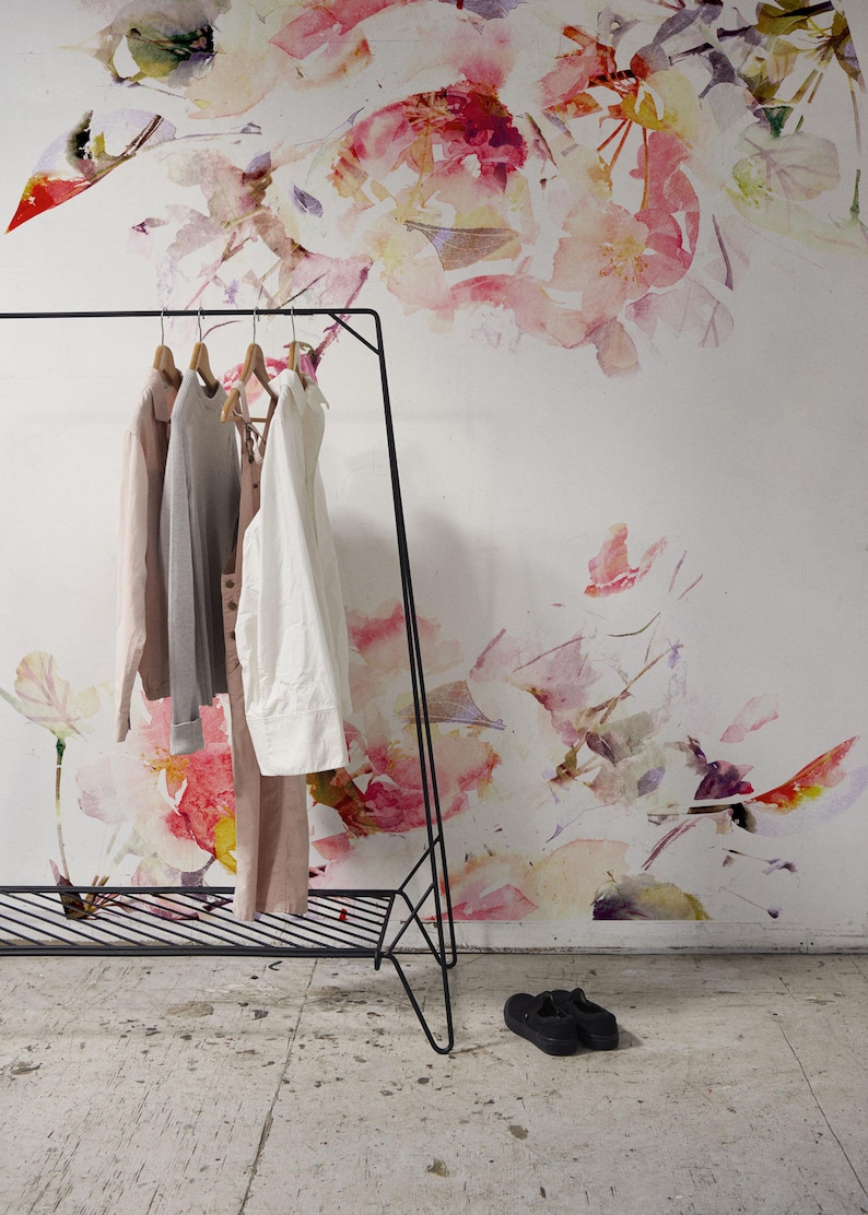 May include: A black metal clothing rack with a shelf and four garments hanging on it. The rack is in front of a white wall with a watercolor floral pattern.