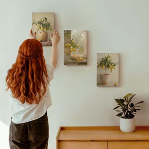 May include: A person hanging three rectangular canvas art prints featuring lemons. The prints show lemon trees in vases and pots, and lemons in a bowl. The art has a vintage aesthetic, with a neutral colour palette.