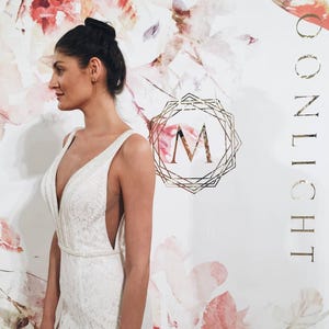 May include: A woman wearing a white lace wedding dress with a plunging neckline and a beaded embellishment. The dress has a fitted bodice and a flowing skirt. The background is a white wall with a floral pattern and the text "MOONLIGHT" in gold lettering.