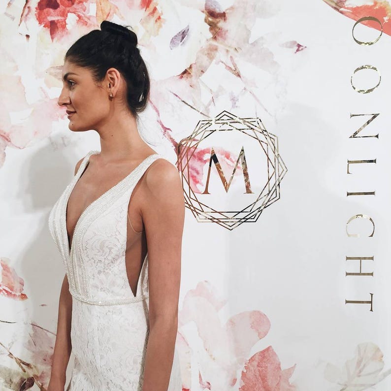 May include: A woman wearing a white lace wedding dress with a plunging neckline and a beaded embellishment. The dress has a fitted bodice and a flowing skirt. The background is a white wall with a floral pattern and the text "MOONLIGHT" in gold lettering.