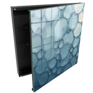 Op de afbeelding: Een wandgemonteerde sleutelkast met een zwart metalen frame en een glazen voorpaneel. Het glas is voorzien van een printontwerp van overlappende cirkelvormige bellen in blauwe, grijze en witte tinten. Het zwarte binnenpaneel bevat vier kleine metalen haken.