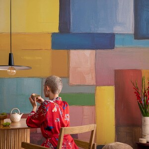 May include: A person in a red floral robe sits at a wooden table, holding a cup. A white teapot and a vase of red flowers are on the table. The background is a colorful abstract painting with rectangular blocks of color.