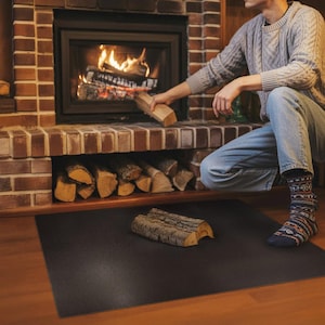 May include: A person adding a log to a burning fireplace. The brick fireplace has a shelf with stacked firewood. A black mat is on the floor with a log on it. The person is wearing a gray sweater and blue jeans.