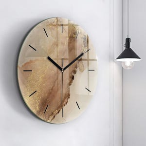 May include: A round wall clock featuring a smooth, glossy surface with an abstract design in shades of beige, cream, and brown, accented by gold flecks. The clock has thin black hour markers and two black hands centered on the face.