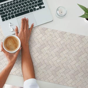 May include: A white ceramic coffee mug with a brown coffee drink inside. The mug is being held by a person's hand. The mug is sitting on a white desk with a light brown patterned desk mat.