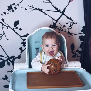 May include: A baby sitting in a high chair with a blue tray. The baby is holding a brown and clear textured ball. The high chair has a white frame and a blue seat. The tray has a brown silicone mat. The high chair has the word "ORIBEL" printed on the front.