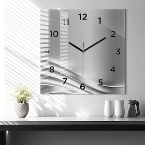 Könnte beinhalten: Eine quadratische Wanduhr mit einer reflektierenden silber-weißen Oberfläche und einem fließenden, abstrakten Wellenmuster. Das Zifferblatt zeigt kräftige schwarze arabische Ziffern von 1 bis 12 und zwei schwarze Zeiger in der Mitte.