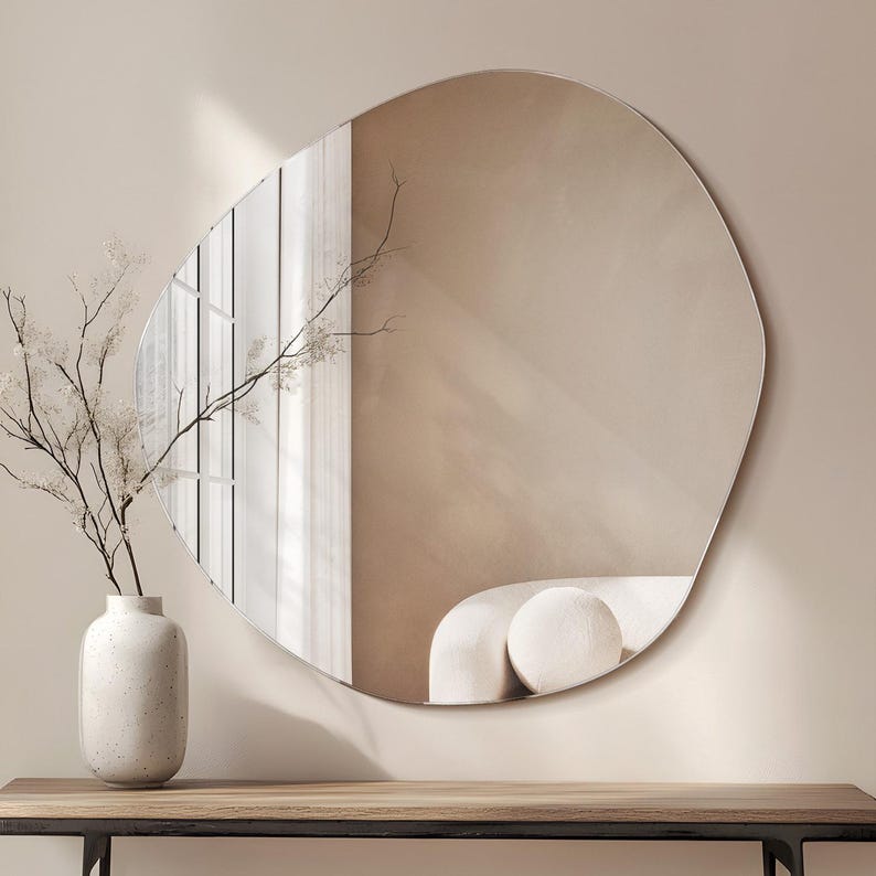 Peut inclure: Un grand miroir mural de forme irr&eacute;guli&egrave;re avec un bord biseaut&eacute;. Le miroir refl&egrave;te une pi&egrave;ce avec des rideaux blancs et un canap&eacute; blanc.