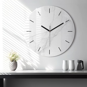 Puede incluir: Un reloj de pared redondo con un patrón de mármol blanco y vetas grises. La esfera del reloj tiene marcadores de hora negros finos y dos manecillas negras. El diseño es limpio y circular, ofreciendo una lectura clara de la hora.