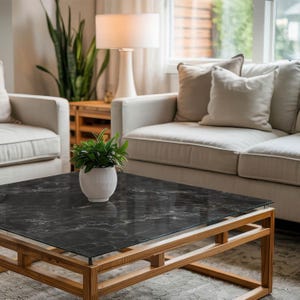 Könnte beinhalten: Ein modernes Wohnzimmer mit einem quadratischen Couchtisch. Der Tisch hat eine schwarze Marmoroptik-Oberfläche und einen hellen Holzrahmen. Ein weißer Keramiktopf mit einer grünen Pflanze steht auf dem Tisch. Ein beiges Sofa, zwei Sessel und eine Lampe sind ebenfalls zu sehen.