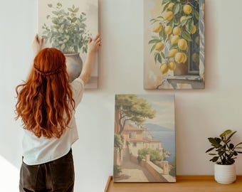 May include: Three canvas art prints displayed on a white wall. The prints depict a vase of greenery, a lemon tree, and a coastal landscape. A person with red hair is hanging the first print. A wooden cabinet is below the prints.