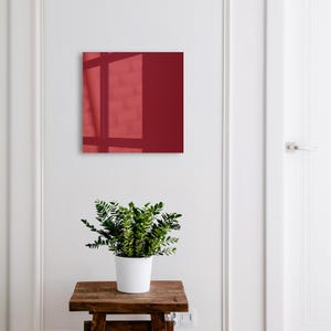 Può includere: Un'opera d'arte da parete quadrata di colore rosso intenso con una superficie lucida e riflettente che mostra il tenue riflesso di una finestra. L'opera è montata su una parete bianca sopra uno sgabello in legno con una pianta verde.