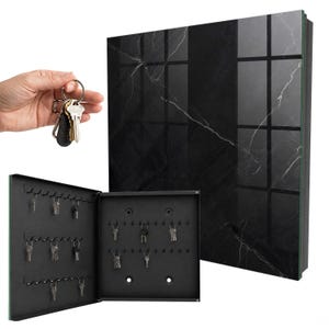 Può includere: Un armadietto portachiavi da parete nero con un pannello frontale lucido con motivo in marmo nero. L'armadietto si apre per rivelare un interno nero opaco con diverse file di ganci metallici per appendere le chiavi. È mostrato un mazzo di chiavi.
