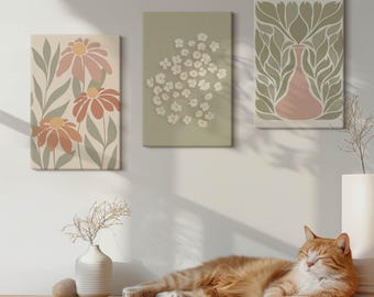 Può includere: Tre stampe artistiche floreali su tela nei toni tenui del rosa, verde e beige sono appese a una parete bianca. Un gatto rosso si riposa su una superficie di legno davanti a un vaso con fiori secchi. L'arte presenta fiori e foglie stilizzate.