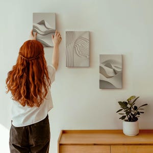 May include: Three abstract art canvas prints in shades of gray and white are displayed on a white wall. One print is being hung by a person with long red hair. The prints feature layered, textured designs reminiscent of landscapes.