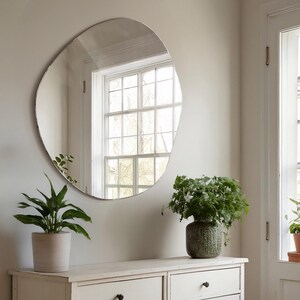 Peut inclure: Une commode blanche &agrave; deux tiroirs surmont&eacute;e d'un grand miroir de forme organique. Le miroir refl&egrave;te une fen&ecirc;tre donnant sur des arbres &agrave; l'ext&eacute;rieur. Deux plantes en pot sont pos&eacute;es sur la commode, l'une avec des feuilles vertes et l'autre avec un pot &agrave; motifs verts et blancs.