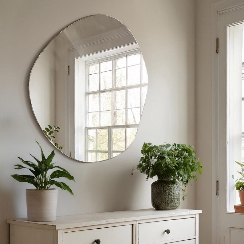 Peut inclure: Une commode blanche &agrave; deux tiroirs surmont&eacute;e d'un grand miroir de forme organique. Le miroir refl&egrave;te une fen&ecirc;tre donnant sur des arbres &agrave; l'ext&eacute;rieur. Deux plantes en pot sont pos&eacute;es sur la commode, l'une avec des feuilles vertes et l'autre avec un pot &agrave; motifs verts et blancs.