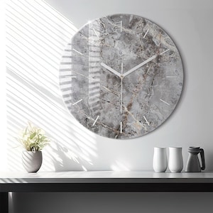 Può includere: Un orologio da parete circolare con un motivo in marmo grigio e bianco sul quadrante. L'orologio presenta sottili indicatori bianchi per le ore e due lancette bianche sottili. La superficie ha una finitura lucida che riflette la luce.