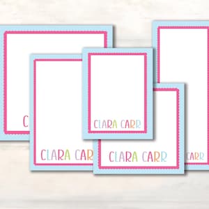 May include: A set of five rectangular notepads in varying sizes, each featuring a white center, a light blue border, and a scalloped hot pink frame. The name Clara Carr is printed in multicolored letters on the bottom of four of the notepads.
