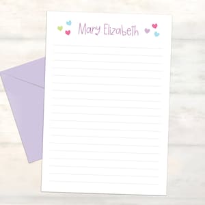 May include: White lined stationery with the name "Mary Elizabeth" in purple script, decorated with small hearts in shades of pink, green, and blue. A matching lavender envelope is in the background. Stationery for letter writing.