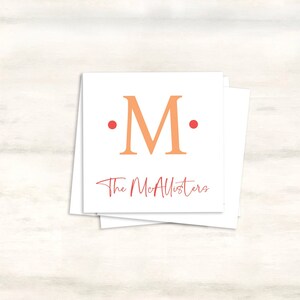 K&ouml;nnte beinhalten: Eine wei&szlig;e Karte mit einem gro&szlig;en orangefarbenen "M" in der Mitte, umgeben von zwei roten Punkten. Der Text "The McAllisters" ist in roter Kursivschrift unter dem Buchstaben geschrieben.