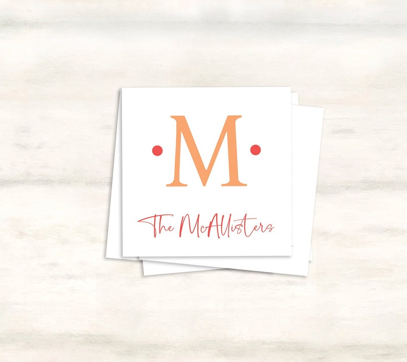 K&ouml;nnte beinhalten: Eine wei&szlig;e Karte mit einem gro&szlig;en orangefarbenen "M" in der Mitte, umgeben von zwei roten Punkten. Der Text "The McAllisters" ist in roter Kursivschrift unter dem Buchstaben geschrieben.