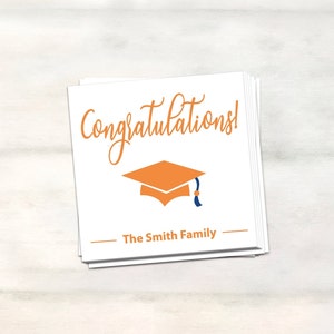 Puede incluir: Una tarjeta blanca con el texto "&iexcl;Felicidades!" en letra naranja. Debajo del texto hay una peque&ntilde;a gorra de graduaci&oacute;n naranja con un borla azul. El texto "The Smith Family" est&aacute; impreso en negro debajo de la gorra.