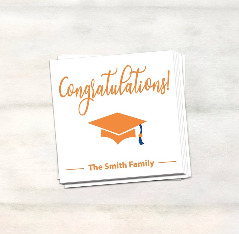 Puede incluir: Una tarjeta blanca con el texto "&iexcl;Felicidades!" en letra naranja. Debajo del texto hay una peque&ntilde;a gorra de graduaci&oacute;n naranja con un borla azul. El texto "The Smith Family" est&aacute; impreso en negro debajo de la gorra.