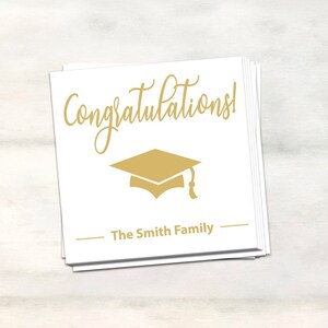 Puede incluir: Tarjeta blanca con texto dorado que dice "&iexcl;Felicidades!" y un icono de gorro de graduaci&oacute;n dorado. La tarjeta tambi&eacute;n tiene el texto "The Smith Family" en dorado.