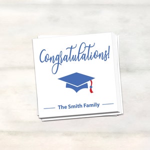 Puede incluir: Tarjeta blanca con texto azul en cursiva que dice "&iexcl;Felicidades!" y un gorro de graduaci&oacute;n azul con una borla roja. La tarjeta tambi&eacute;n tiene una l&iacute;nea con el texto "The Smith Family" impreso debajo del gorro.