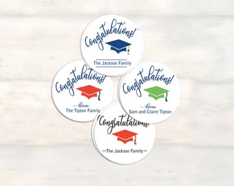 May include: Four round white stickers with the text "Congratulations!" in blue cursive writing. Each sticker features a graduation cap in a different color: blue, red, green, and red. The stickers also include the text "from" and the name of the sender.