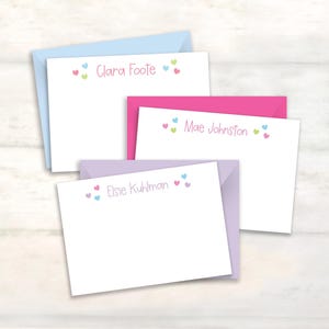 May include: Three white rectangular note cards with matching envelopes in light blue, hot pink, and lavender. Each card features a personalized name printed in pink, flanked by small colorful heart icons in pink, blue, and lime green.