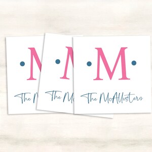 K&ouml;nnte beinhalten: Drei wei&szlig;e Karten mit einem rosa Buchstaben "M" und zwei blauen Punkten auf jeder Karte. Der Text "The McAllisters" ist in blauer Kursivschrift auf der untersten Karte rechts geschrieben.